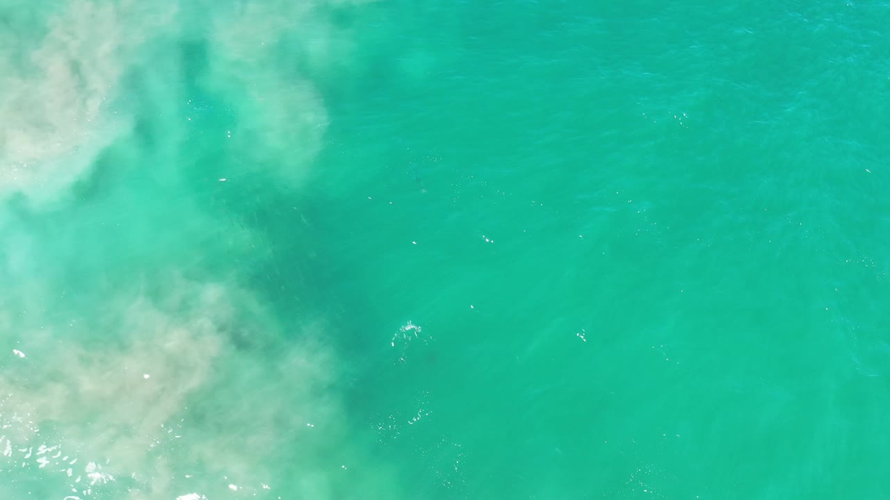 Overhead view of Los Cabos sea water with a thinner foam area near the upper left, scattered surface sparkles, and no visible people.