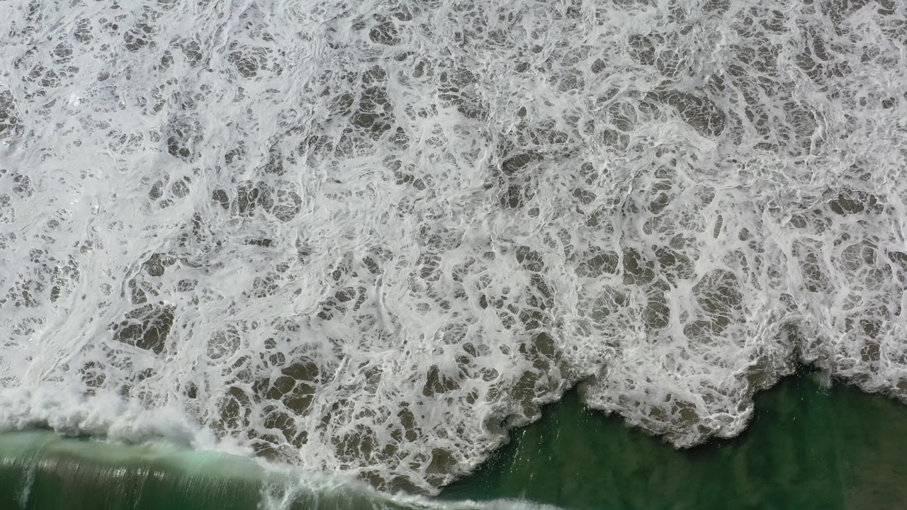 Overhead view in Los Cabos, Mexico, with dense white surf foam spreading over dark green water along the edge of wet sand and no visible people.