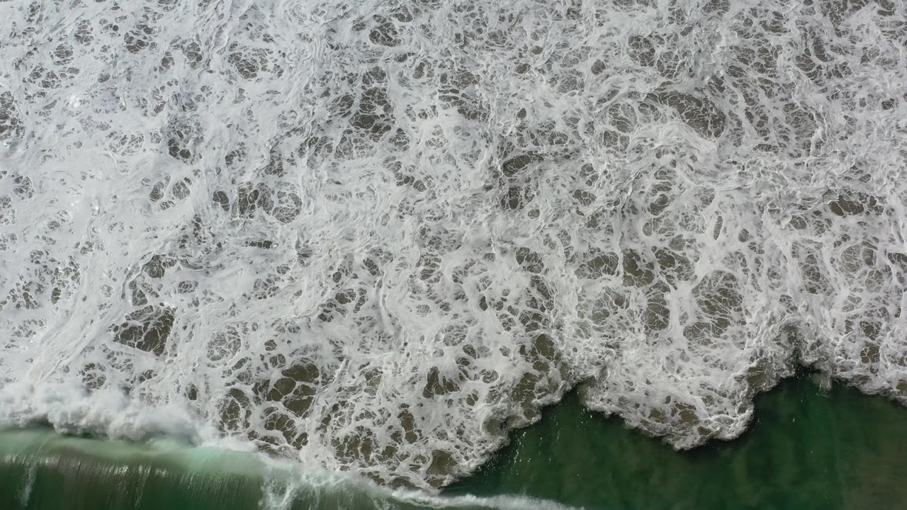Overhead Los Cabos shoreline frame with lace-like white foam covering the upper area, a darker green water band below it, and no visible people.