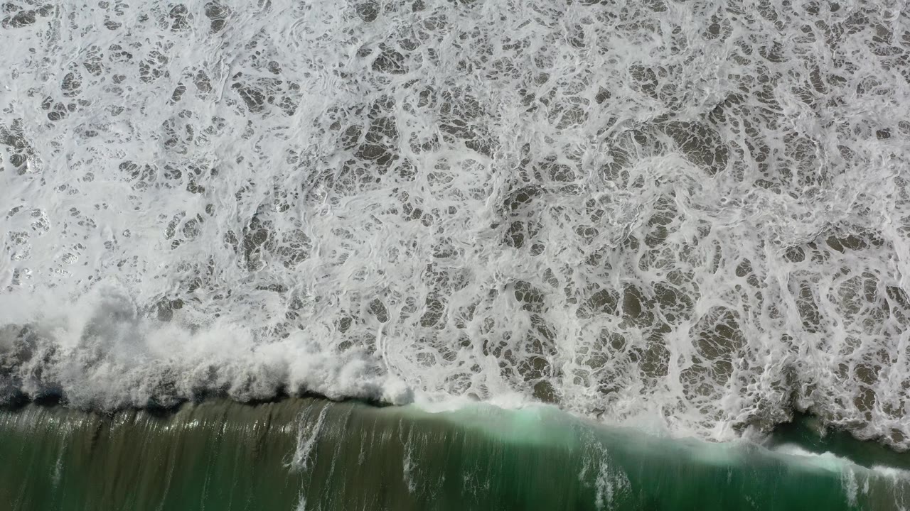 Breaking wave curls across dark green water beneath a broad field of foam.