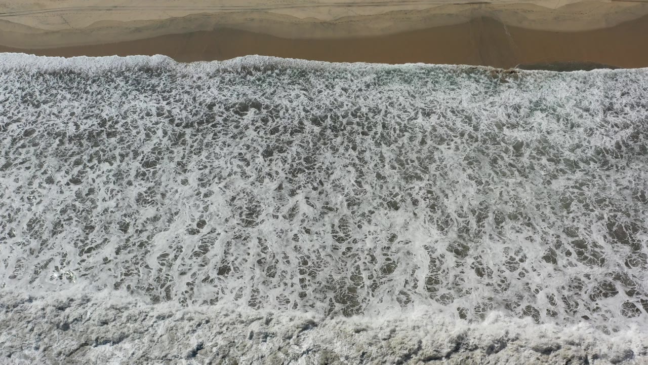 Overhead view of white surf covering the lower edge of a tan Los Cabos beach, with wet sand at the top and no visible people.