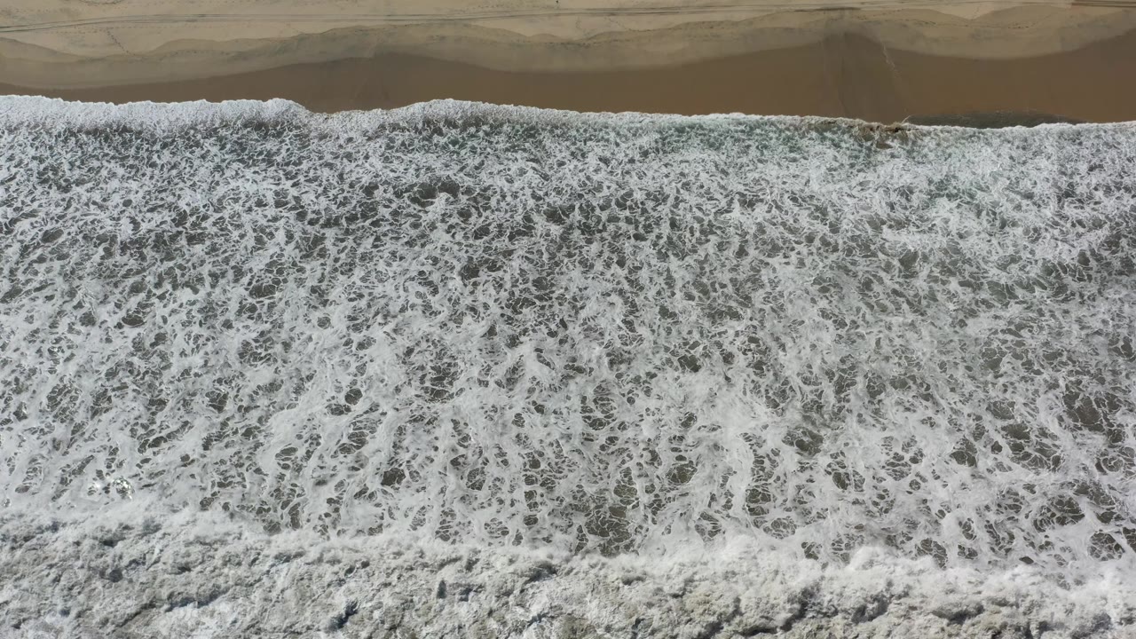 Dense white wave foam fills most of the frame along a Los Cabos shoreline, with a narrow strip of wet tan sand visible above and no visible people.