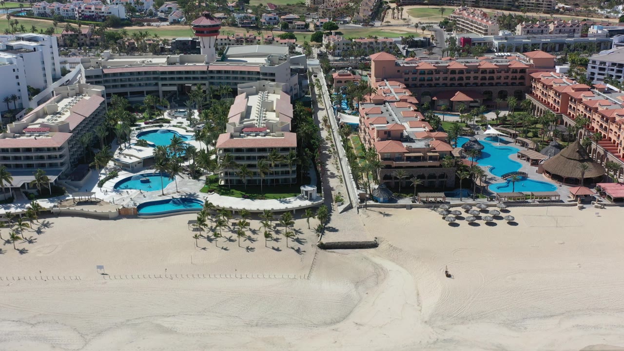 Opening aerial frame over Los Cabos, Mexico, with pale beach sand in front, resort pools and hotel blocks in the middle, suburban buildings beyond, and no visible people.