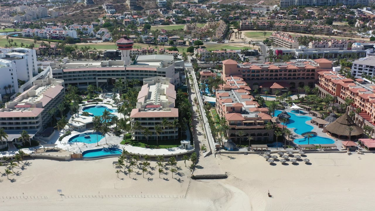 Resort complex with pools beside a wide sandy beach and inland neighborhood.