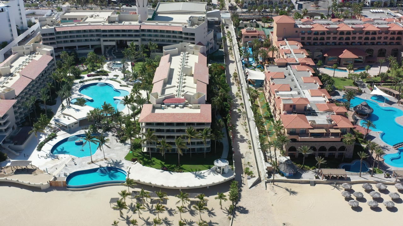 Overhead Los Cabos resort view with a central road or walkway dividing red-roofed buildings, blue pool decks, palm landscaping, beach sand, and no visible people.