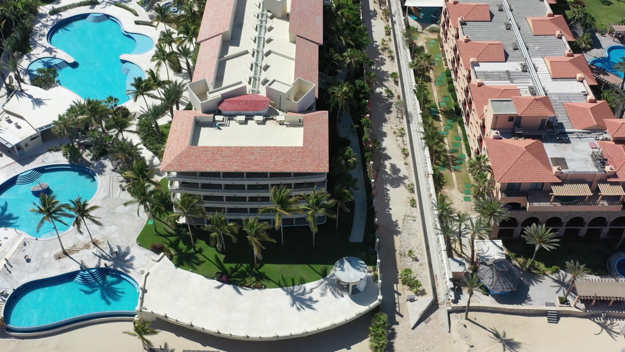 Aerial frame in Los Cabos, Mexico showing red-roof resort structures, a straight narrow road, palms, pool edges, and pale paved resort grounds.