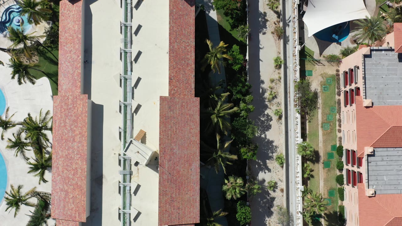 Overhead frame in Los Cabos, Mexico showing red tile roofs, pale roofs, palms, paved paths, shade canopies, and a small pool area with no visible people.