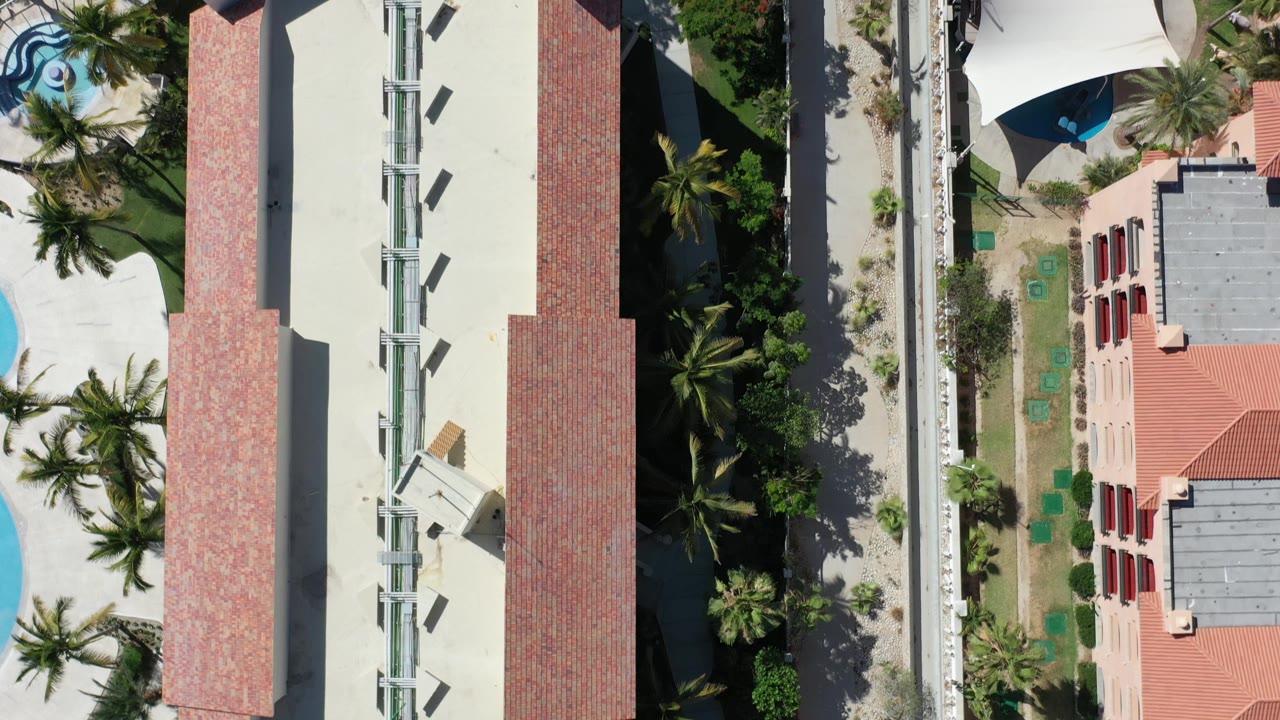 Los Cabos overhead view with red tile roof sections, a long pale roof, palm trees, walkways, planted areas, and no visible people.