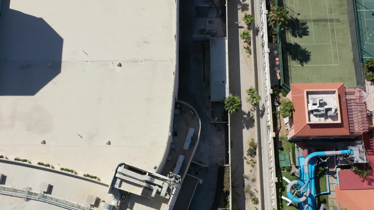 Top-down Los Cabos view of a large pale roof, a green tennis court, palms, and a blue pool slide area, with no visible people.