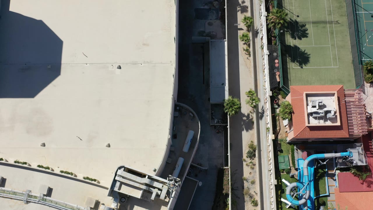 Top-down Los Cabos frame centered on a red-roofed structure near a tennis court, palms, pale roofs, and pool equipment, with no visible people.