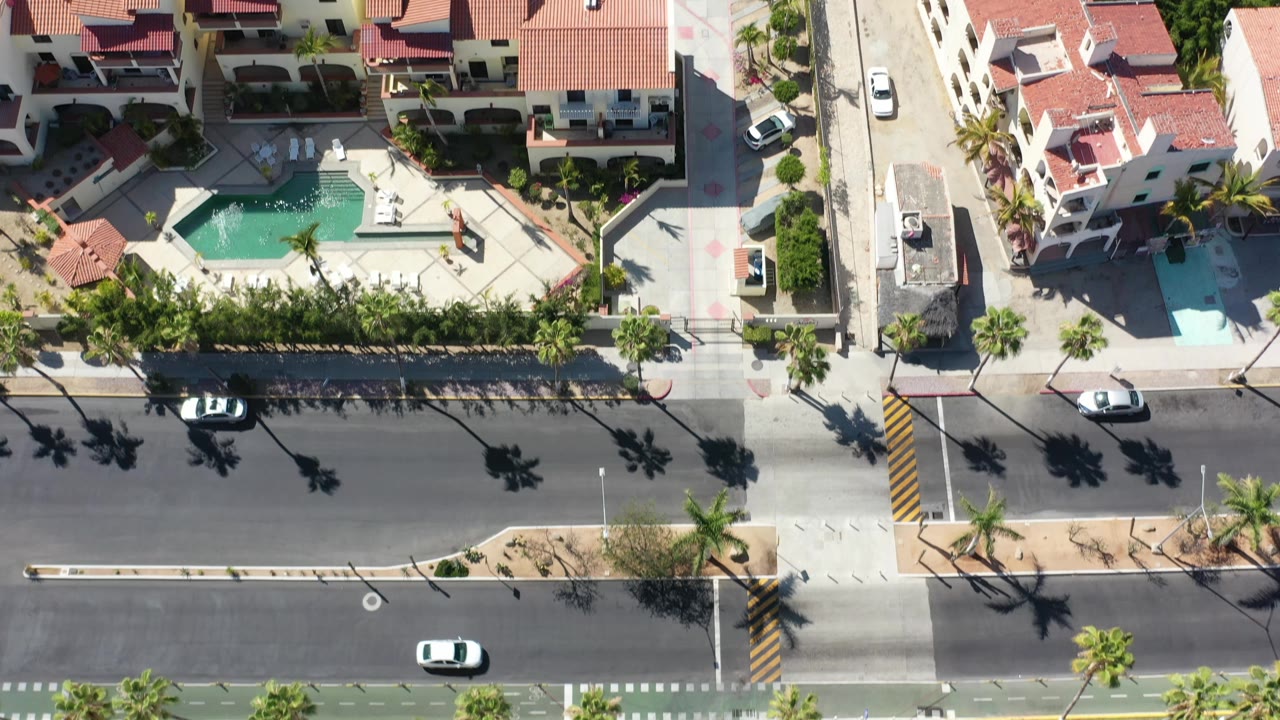 Top-down Los Cabos residential view with red-roof buildings, a turquoise pool courtyard, road markings, cars, palms, and no visible people.