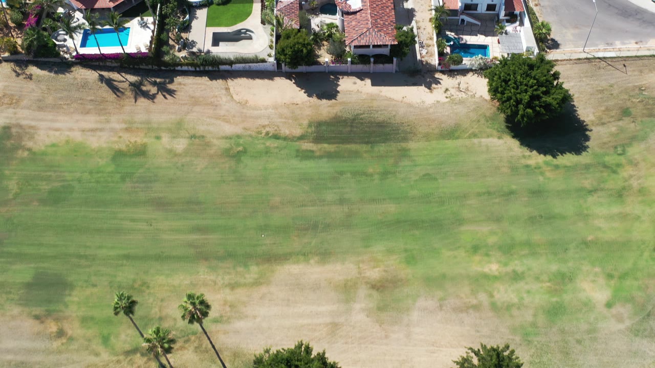 High overhead frame in Los Cabos, Mexico showing patchy green open ground below walled residential properties with tile roofs, palms, and turquoise pools, with no visible people.