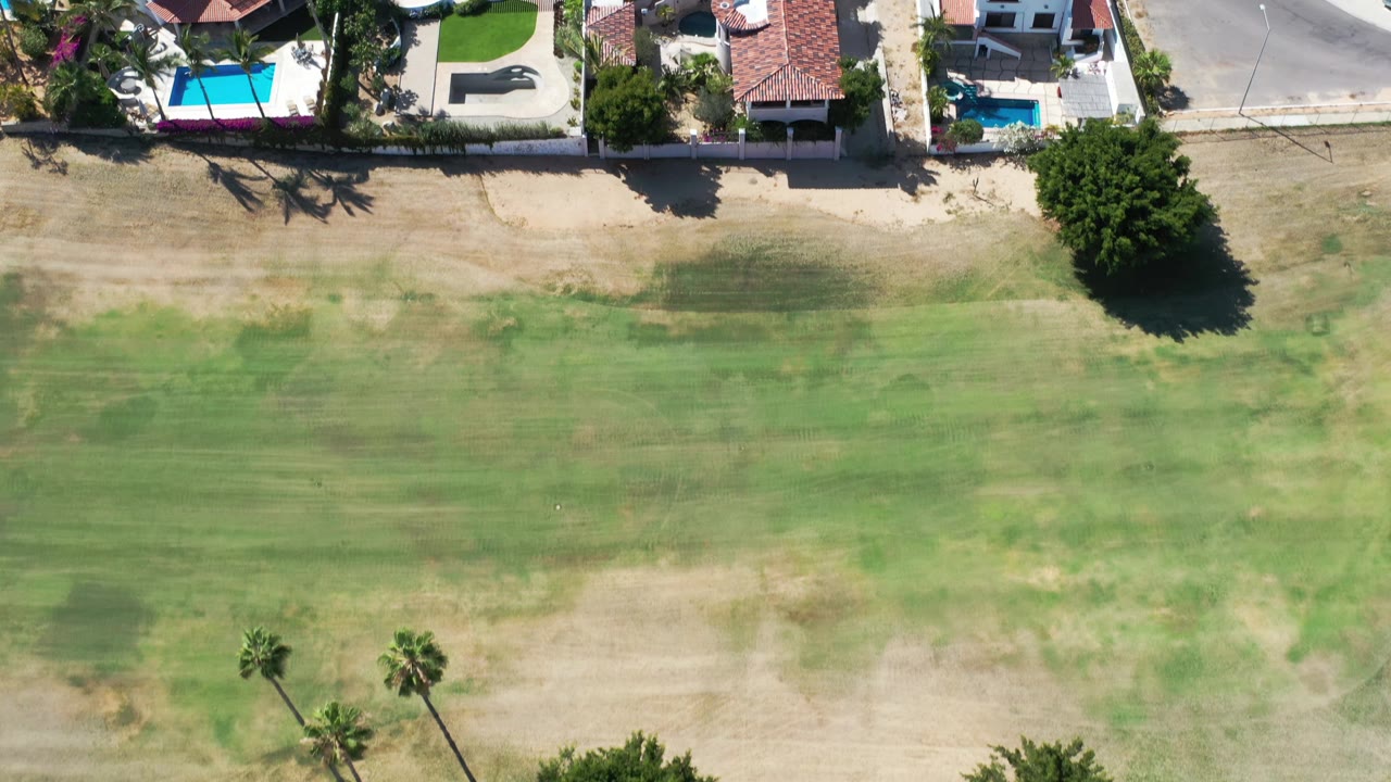 A near-vertical aerial frame shows a broad patch of green and tan ground bordered by Los Cabos homes, boundary walls, small pools, palms, and a road, with no visible people.