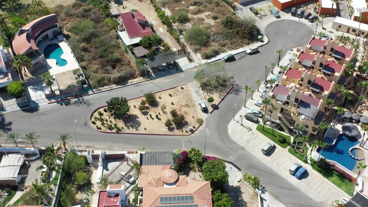 Aerial view in Los Cabos, Mexico with a street junction, cream and red-roofed residences, bright blue pools, dry lots, parked vehicles, and no visible people.