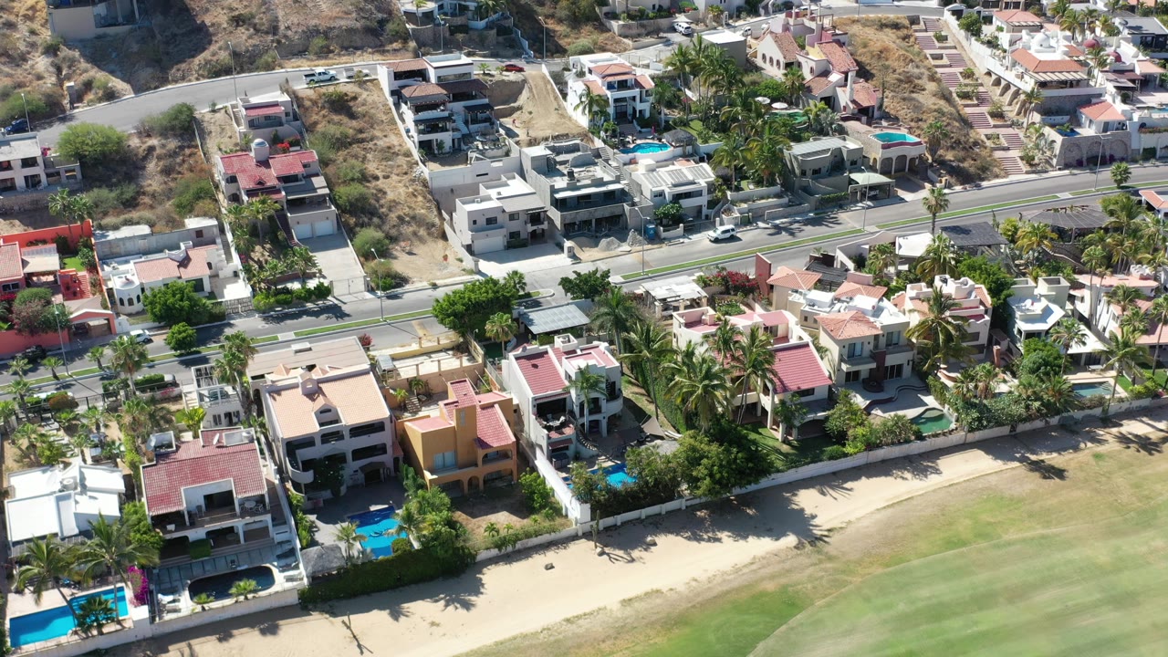 Los Cabos houses with flat and tiled roofs fill the foreground, with turquoise pools, palm landscaping, a two-lane road, hillside homes, and no visible people.