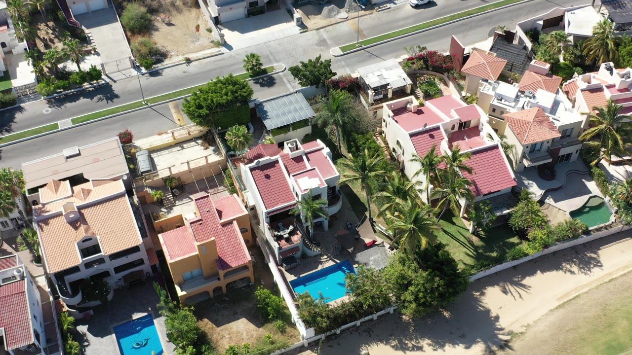 Overhead view of homes with red roofs, pools, palms, and a street.
