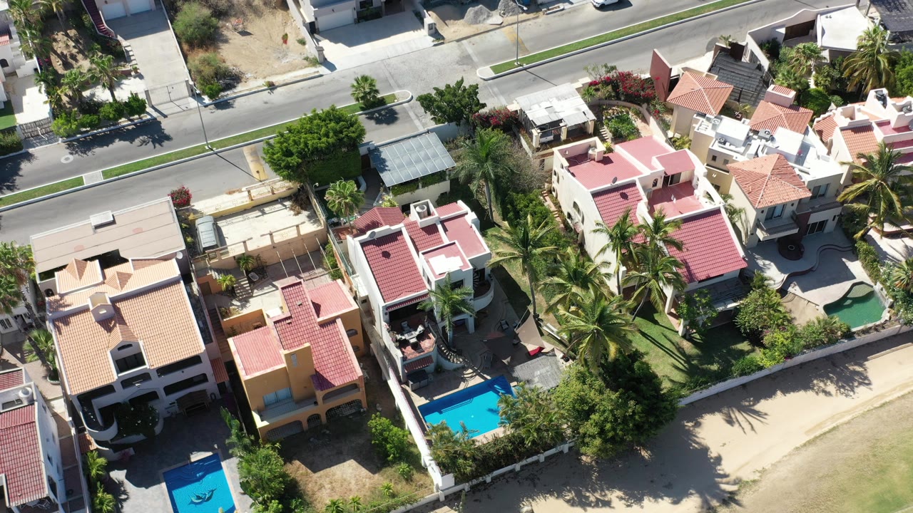 Overhead Los Cabos frame showing clustered private homes, bright pool water, palms along property lines, a paved road, and no visible people.