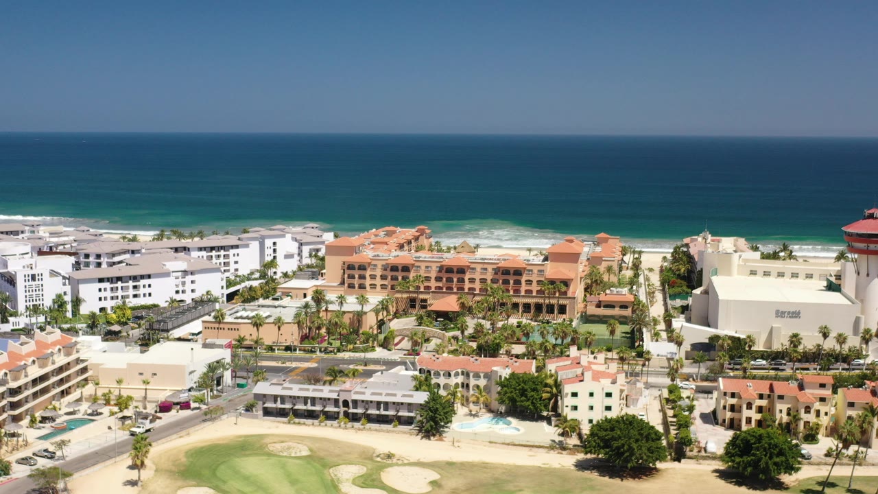 Opening aerial frame over Los Cabos, Mexico with white and terracotta hotel buildings, roads, surf, deep blue ocean, and no visible people.