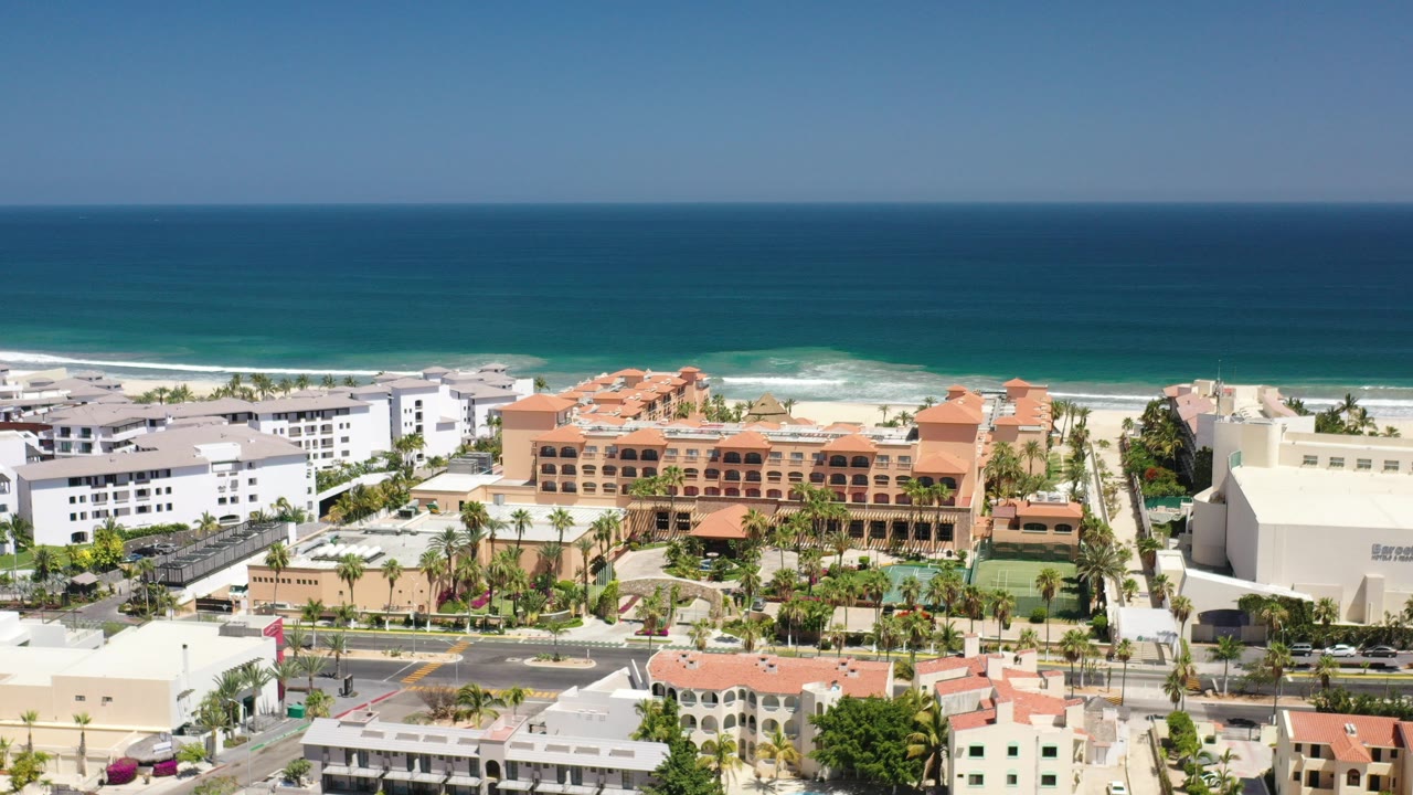 Aerial frame over Los Cabos, Mexico, showing terracotta-roof resort buildings, nearby white hotel blocks, a road, palms, beach, and a broad blue ocean horizon with no clearly visible people.