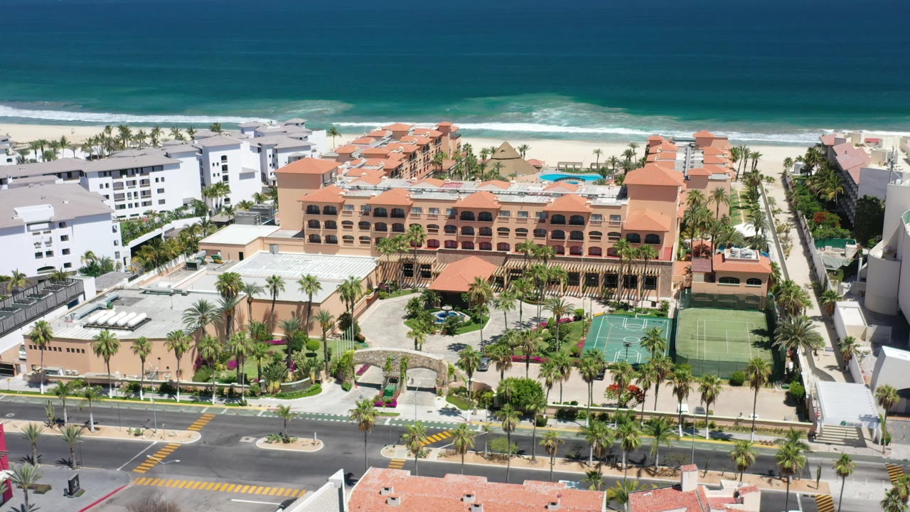 Early aerial frame in Los Cabos, Mexico, with the hotel complex centered between a foreground road and the bright beach and surf line, with no visible people.