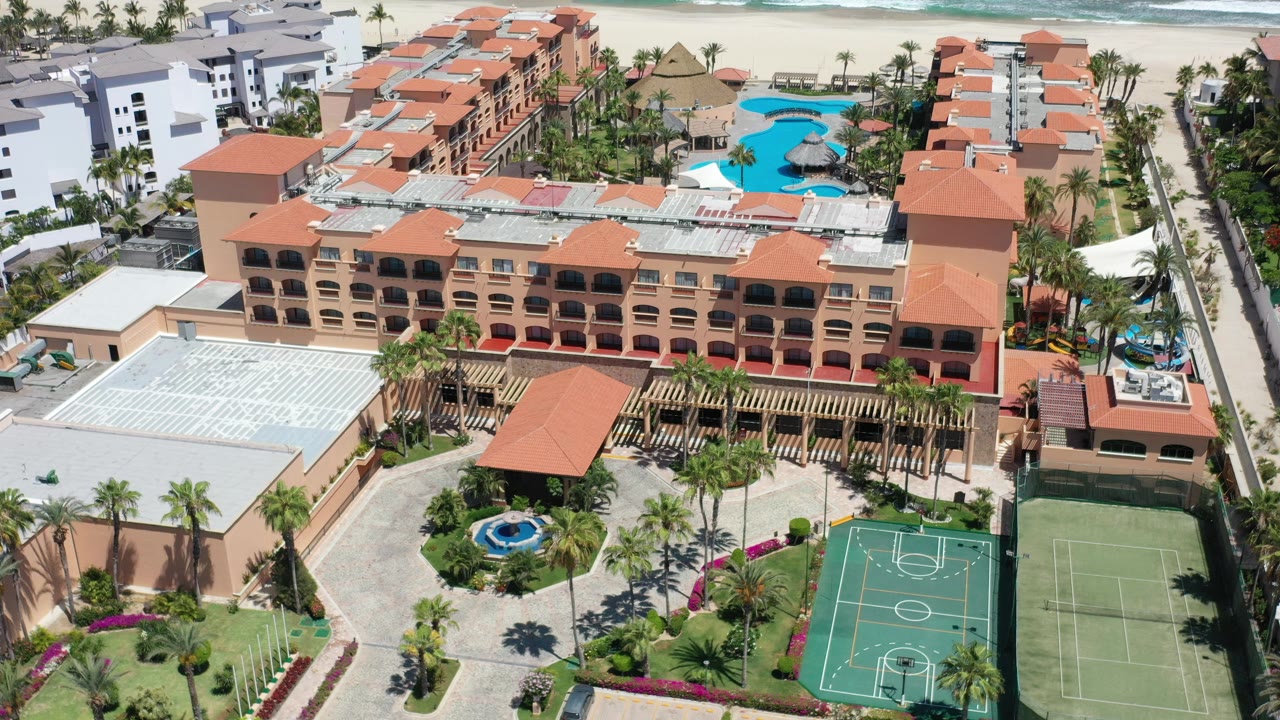Aerial view of terracotta-roofed resort buildings, pools, courts, palms, beach, and ocean.