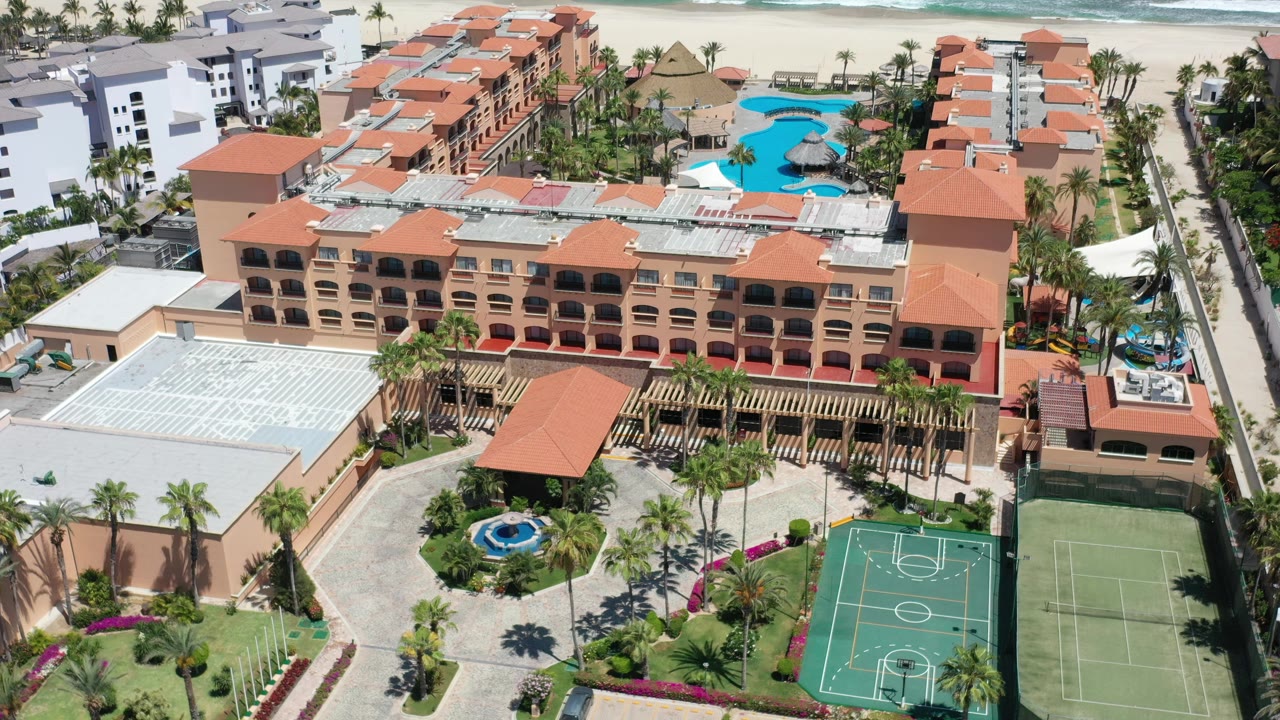High-angle Los Cabos resort frame showing orange roof sections, a paved courtyard with fountain, adjacent green courts, bright pools behind the buildings, beach sand, and no visible people.