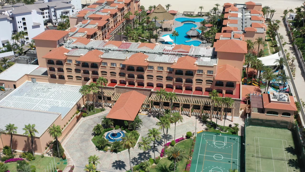 Drone view of resort rooftops, palm-lined grounds, sports courts, pools, and shoreline.