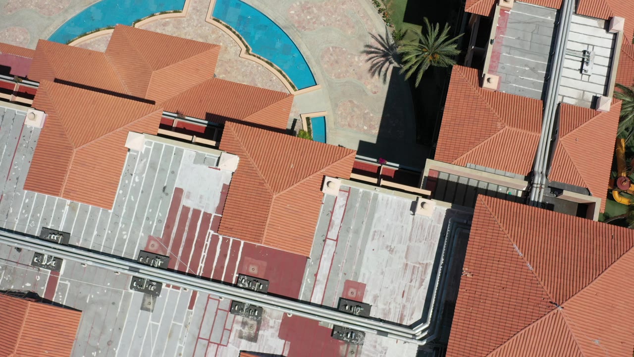 Overhead Los Cabos hotel view with terracotta roofs, pale rooftops, a curved turquoise water feature, palm shadows, and no visible people.