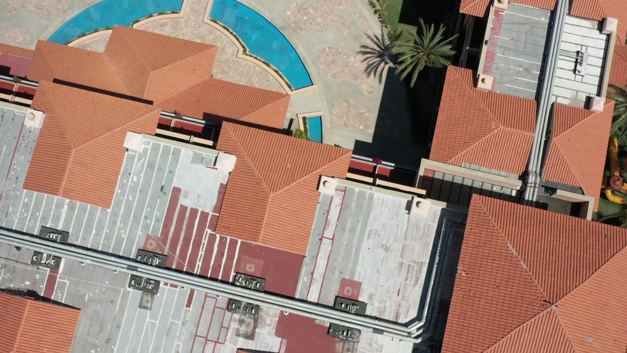 Top-down Los Cabos resort frame showing orange tile roofs, mechanical-looking roof areas, a curved blue pool segment, paved ground, and no visible people.