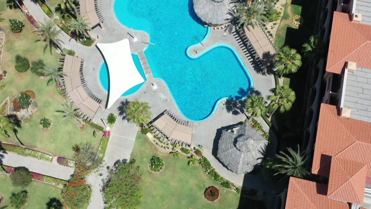 Top-down aerial frame in Los Cabos showing a turquoise pool beside a white shade sail, landscaped grass, palms, paving, and red-roofed buildings, with no visible people.