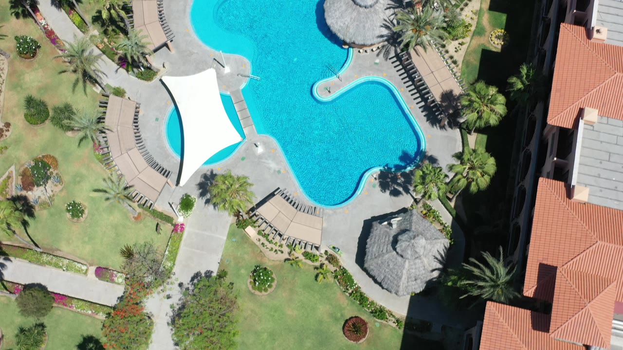 Aerial Los Cabos frame with the curving blue pool centered near a white canopy, lawns and palms to the left, and red-roofed buildings along the right edge, with no visible people.