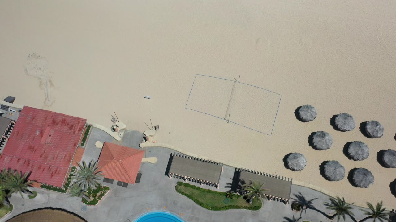 Overhead aerial frame in Los Cabos, Mexico showing pale sand beside red-roofed beachfront structures, palm trees, round thatched umbrellas, and no visible people.