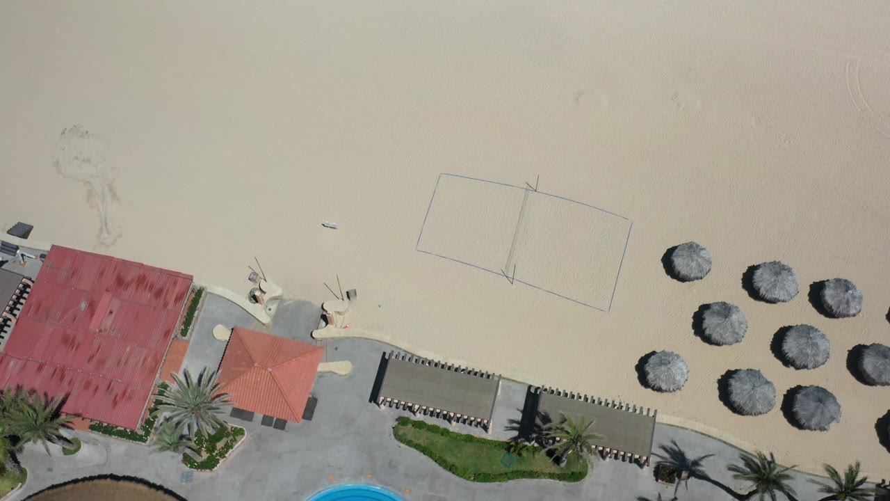 Top-down Los Cabos beach view with red roofing near the lower edge, clustered thatched umbrellas on open sand, faint court markings, and no visible people.