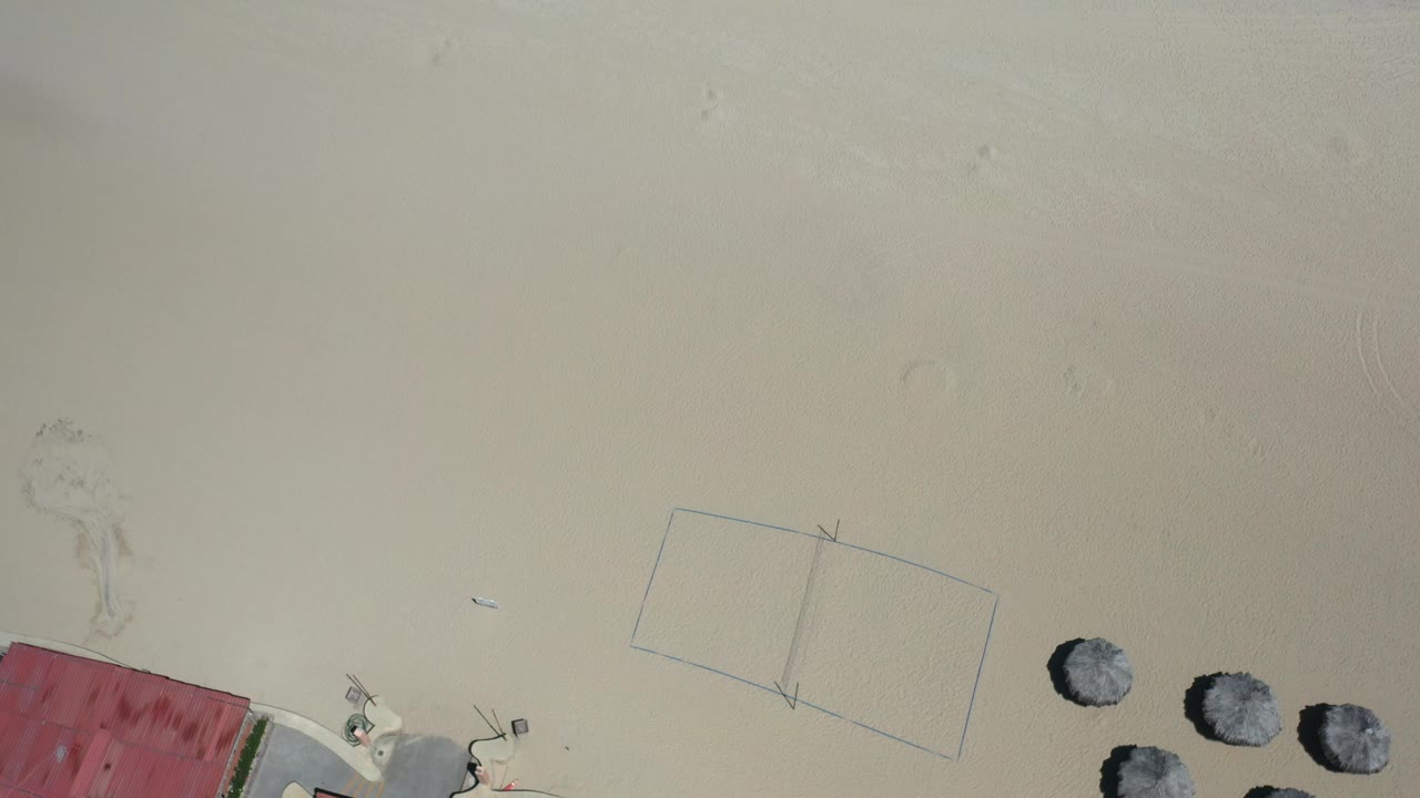 High aerial view of empty sand, court markings, umbrellas, and a red-roofed corner.