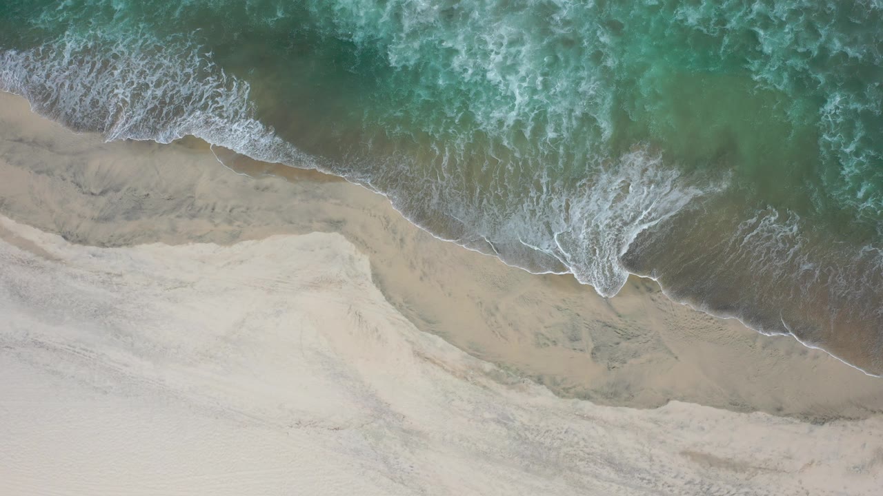 Straight overhead view in Los Cabos, Mexico of green water and white foam meeting pale sand, with no visible people.