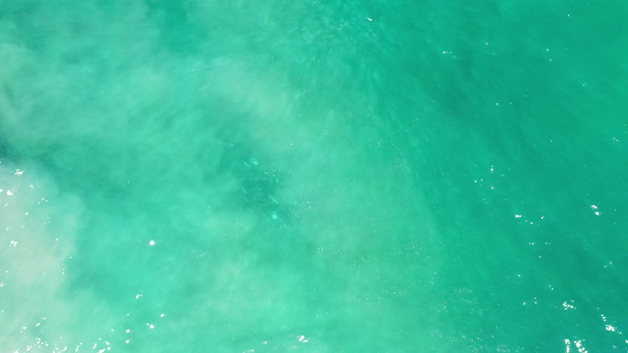 Overhead view of turquoise water in Los Cabos, Mexico, with pale milky surf entering from the left edge and no visible people.