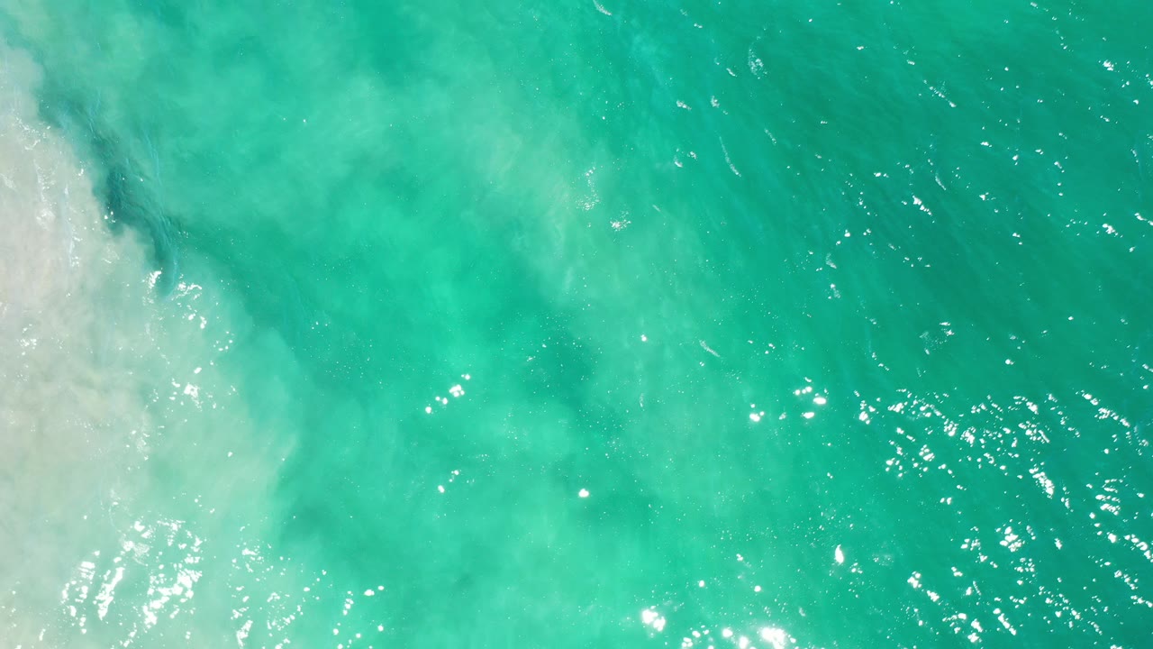 Overhead view of turquoise green sea in Los Cabos, Mexico, with pale foamy surf entering from the left and no visible people.