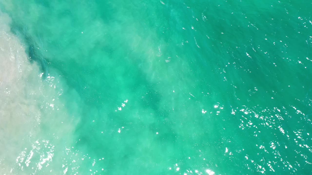 Aerial Los Cabos water view with green-blue ripples, bright sun reflections, and a pale foamy wash along the left side, with no visible people.
