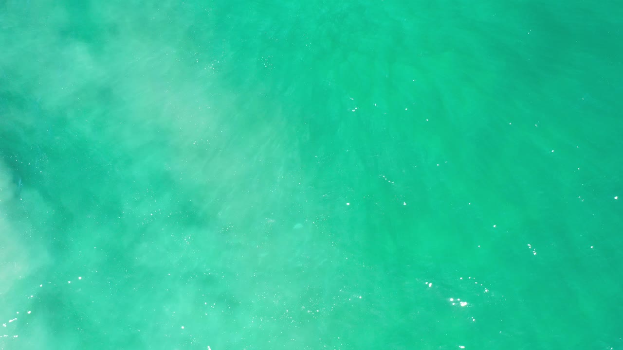 Top-down view of turquoise-green open sea in Los Cabos, Mexico, with pale cloudy water patterns, small sun glints, and no visible people.