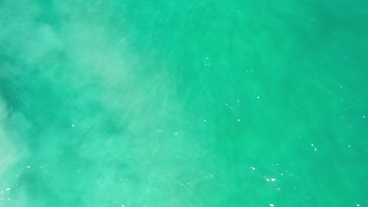 Overhead Los Cabos sea view with green water, milky pale swirls near the center, scattered bright reflections, and no visible people.