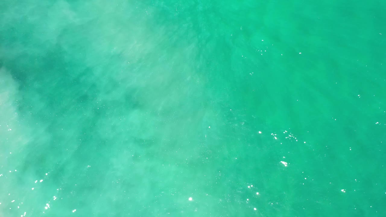 Overhead ocean surface in Los Cabos, Mexico, showing cloudy white wave patterns spread across turquoise water with no visible people.