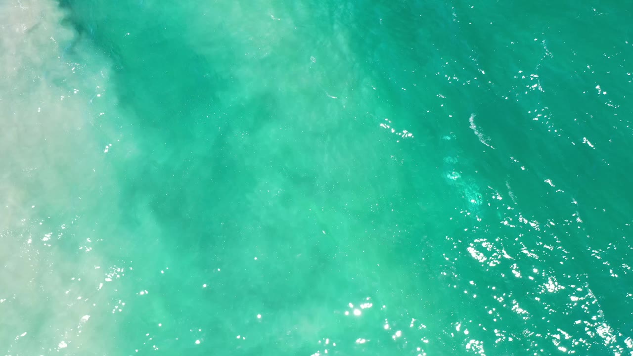 Turquoise water fills the frame in Los Cabos, Mexico, while milky surf foam spreads from the left and small sun highlights speckle the surface, with no visible people.