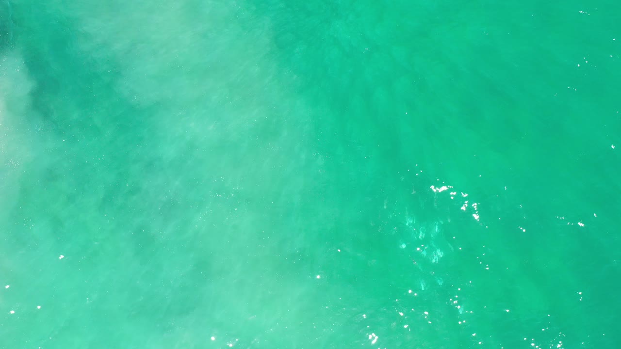 Overhead view of green ocean water in Los Cabos, Mexico, with pale cloudy sand or foam patterns and no visible people.