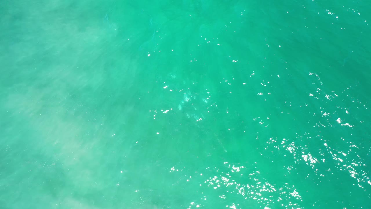 Overhead Los Cabos water view with turquoise color, clustered light reflections near the lower-left area, soft pale patches beneath the surface, and no visible people.