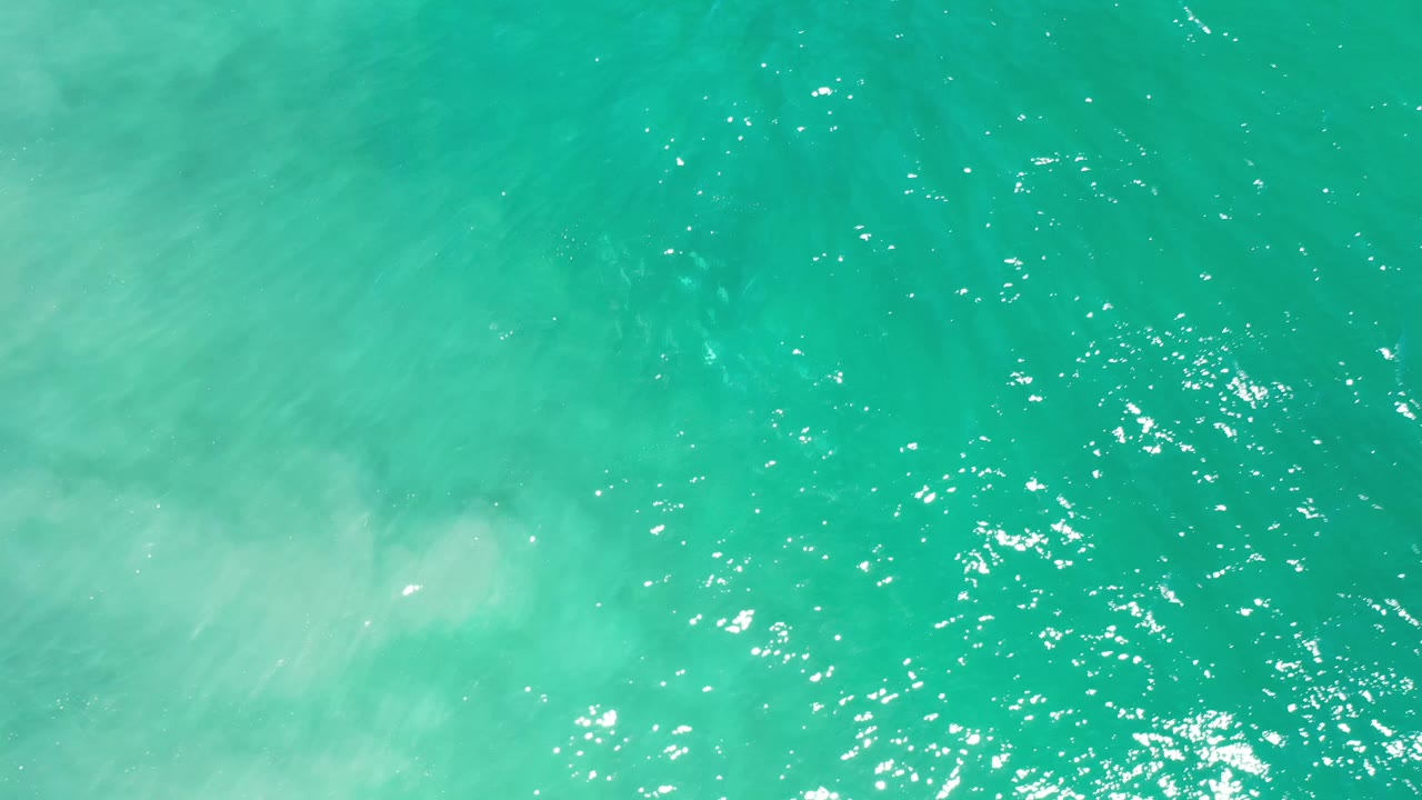 Overhead Los Cabos ocean water with a pale cloudy patch drifting through the blue-green surface and no visible people.