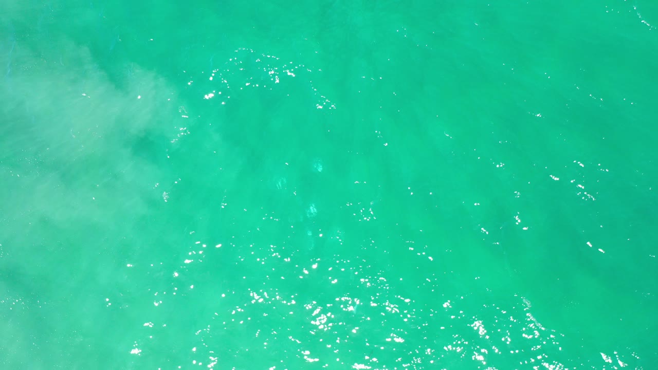 Overhead view of turquoise sea water in Los Cabos with scattered white foam near the upper left and no visible people.