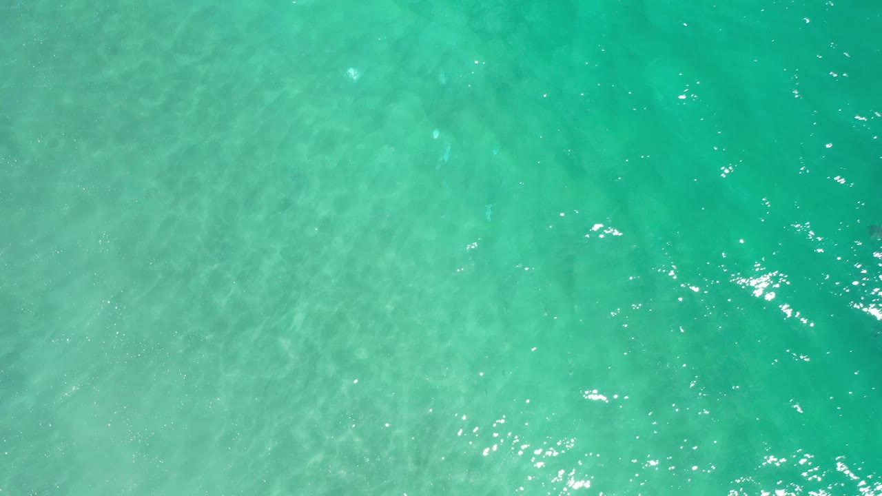 Aerial view of blue-green Los Cabos ocean surface with faint underwater texture, scattered white reflections, and no visible people.