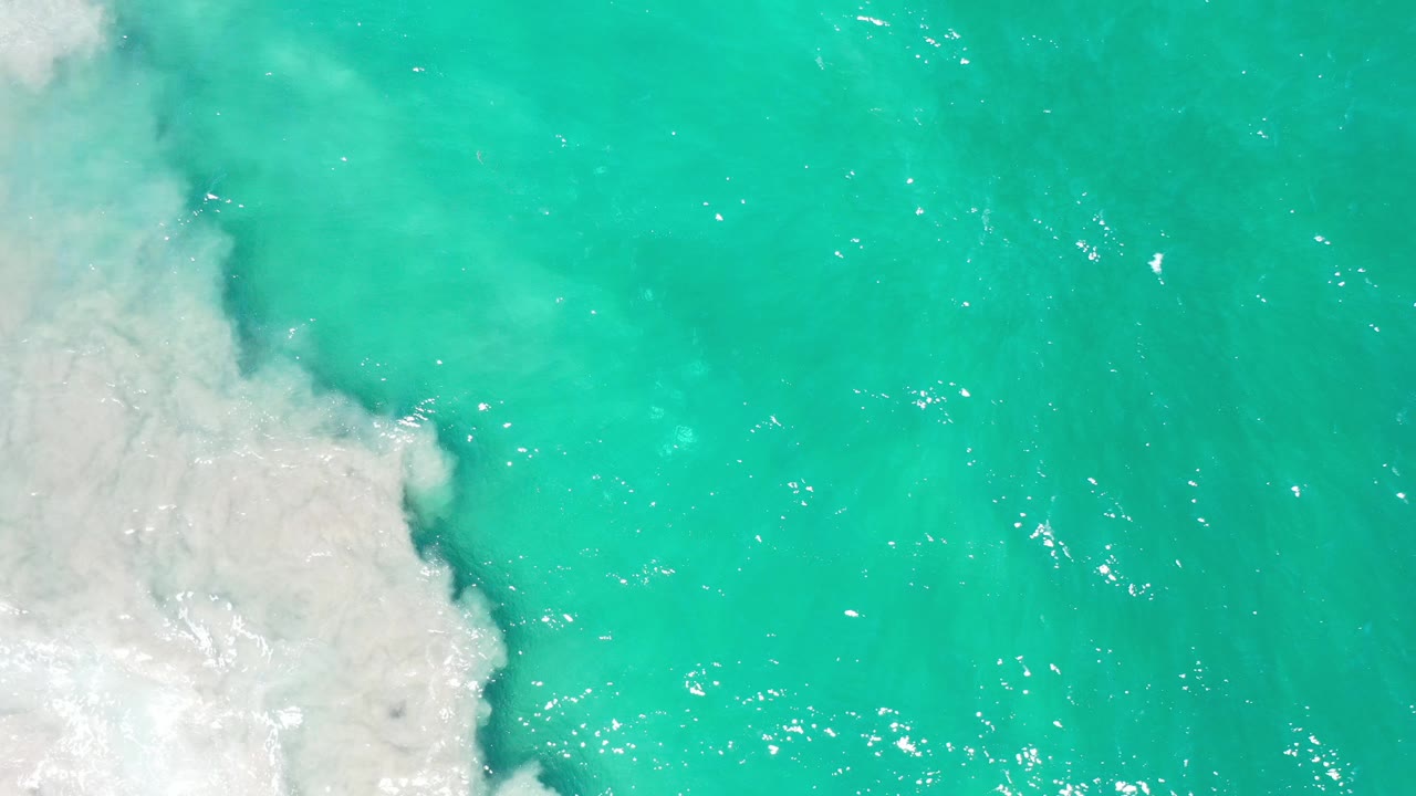 Overhead view of teal-blue water in Los Cabos with pale surf pressing in from the left edge and no visible people.