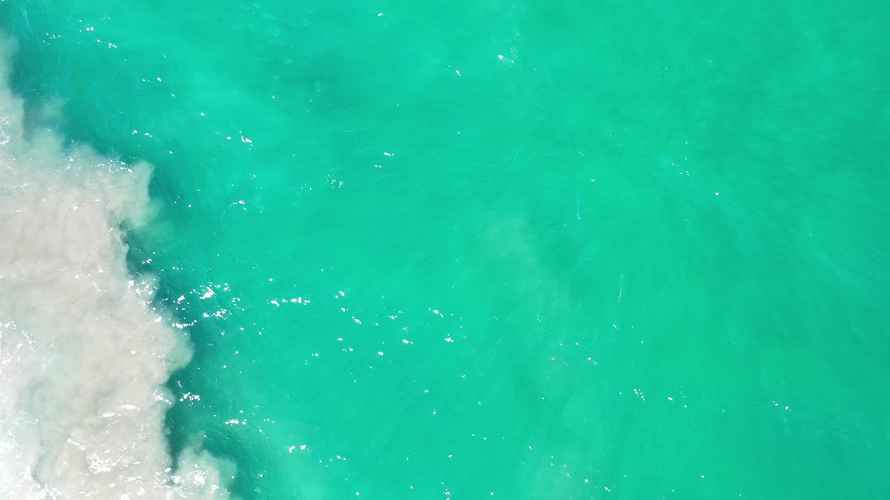 Overhead view of turquoise seawater in Los Cabos, Mexico, with white wave foam pushing in from the left edge and no visible people.