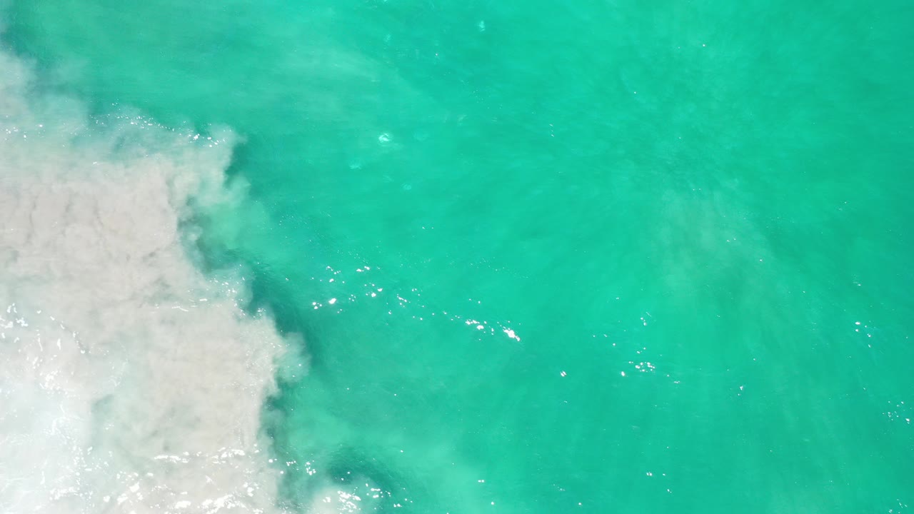 Overhead view of turquoise water in Los Cabos, Mexico, with white foam pushing in from the left side and no visible people.